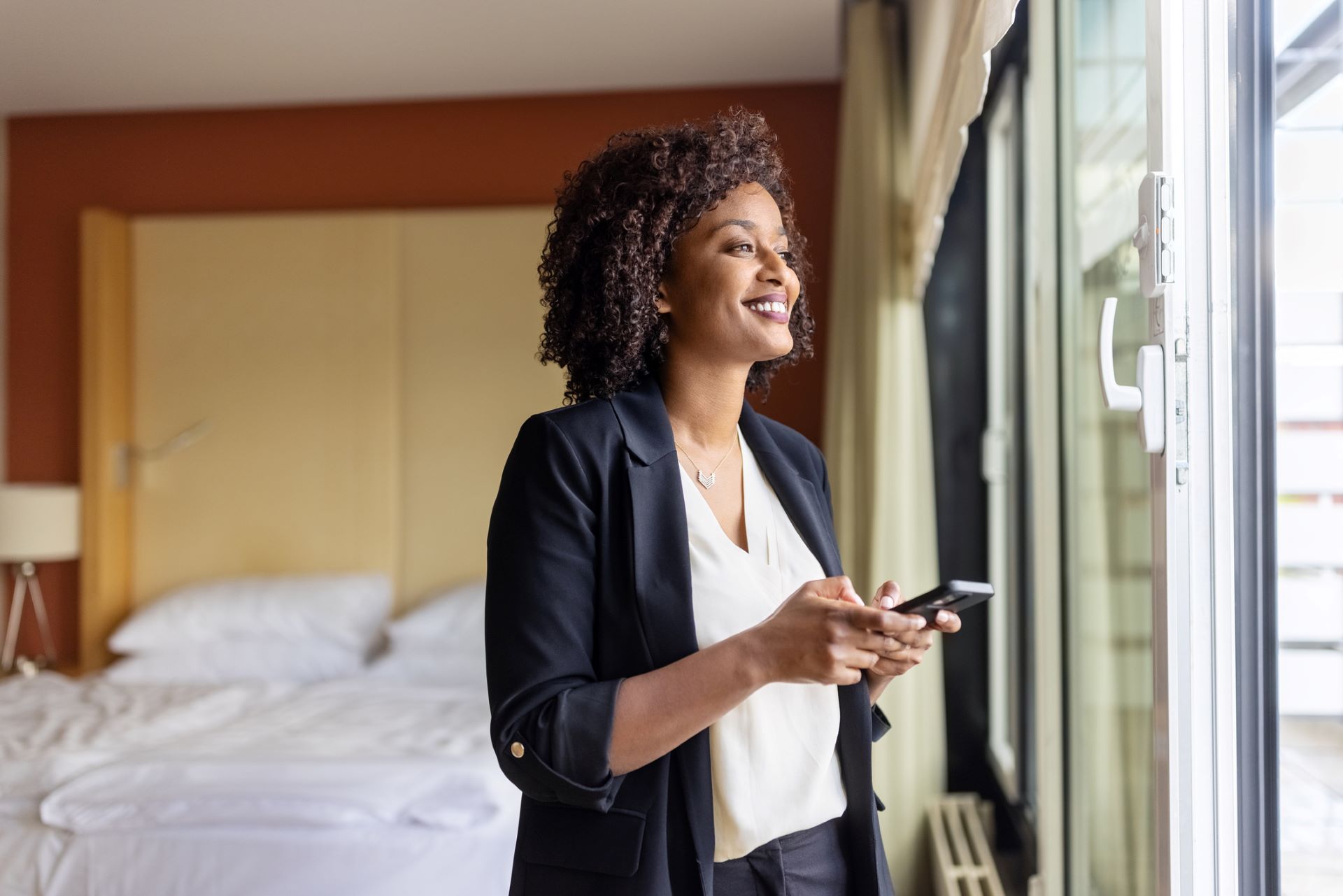 Entspannte Geschäftsfrau im Hotelzimmer am Fenster mit Mobiltelefon in der Hand