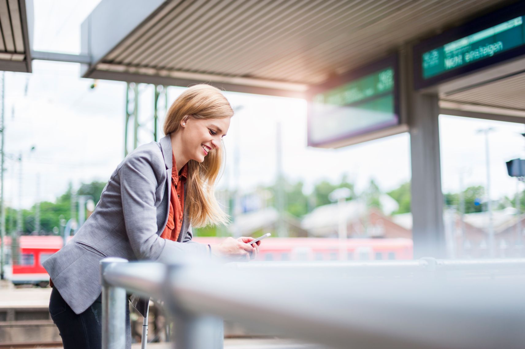 Geschäftsfrau steht lächelnd am Bahnhof und schaut auf ihr Smartphone